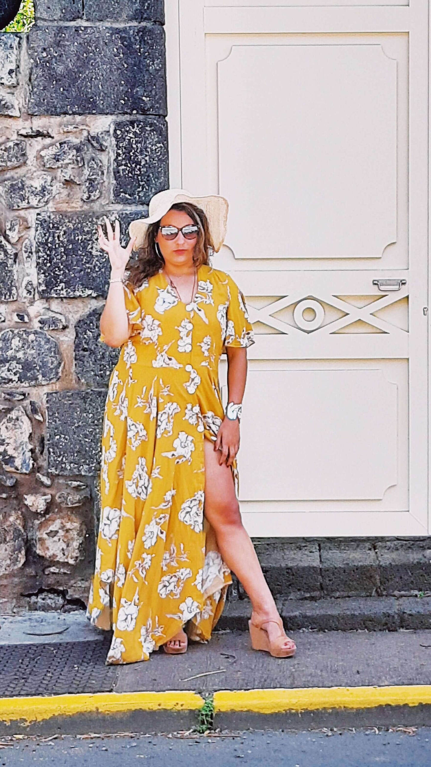 Femme portant une robe longue jaune à fleurs devant une porte, avec un chapeau et des lunettes de soleil.