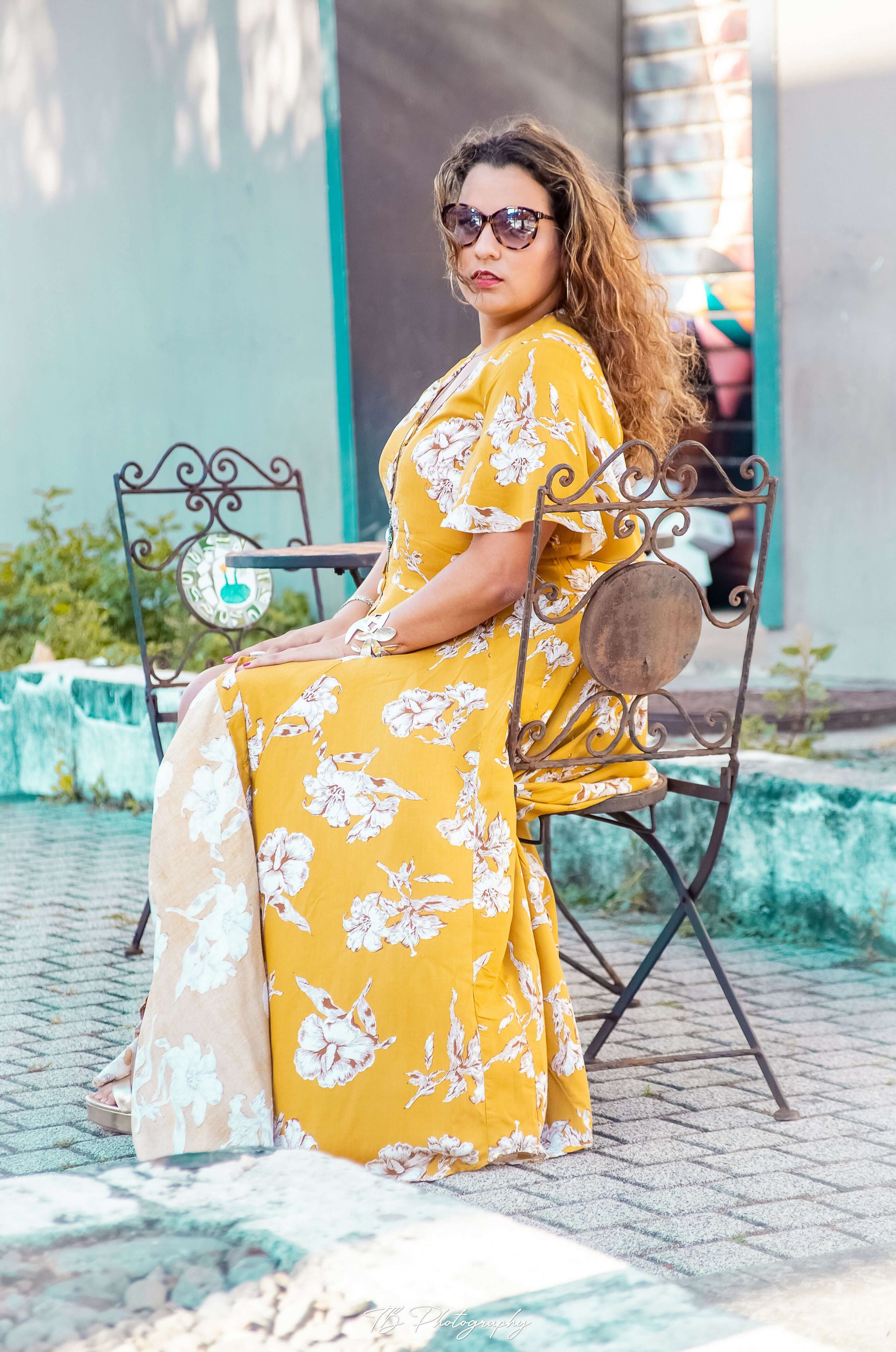 Femme portant une robe longue Aédé à fleurs jaunes, assise sur une chaise en extérieur, mode été chic.