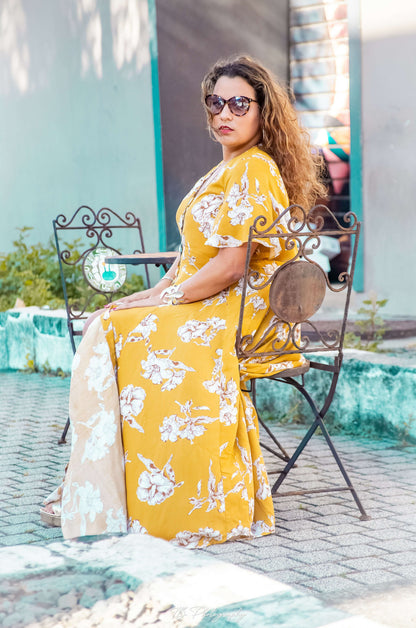 Femme portant une robe longue Aédé à fleurs jaunes, assise sur une chaise en extérieur, mode été chic.