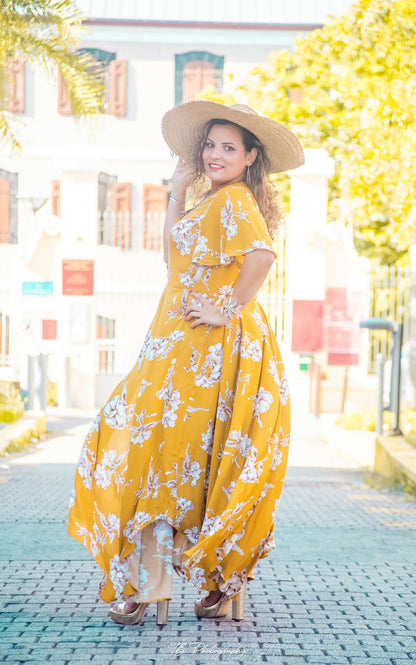Femme souriante portant une robe longue jaune floral avec chapeau, posant en plein air, ambiance estivale.