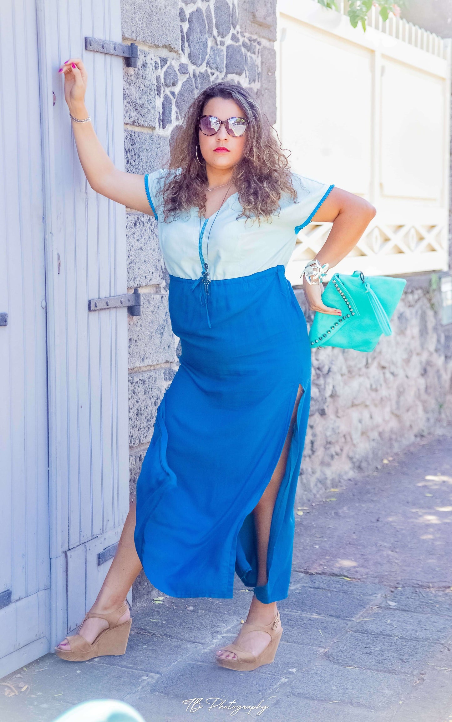 Femme portant une robe longue bleue avec fentes, appuyée contre un mur en pierre, accessoirisée avec un sac turquoise et des sandales compensées.
