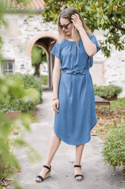 Femme en robe FLORISSE bleu, ceinturée, avec décolleté et manches volantées, dans un cadre naturel.
