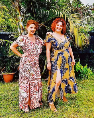 Deux femmes portant des robes longues imprimées posent devant des plantes tropicales.