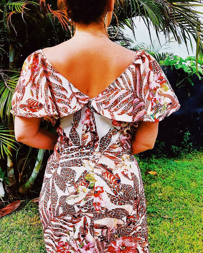 Femme portant la Robe Marpésia - T36 à 50 avec imprimé floral, vue de dos montrant le détail du col en V volanté, jardin en arrière-plan.