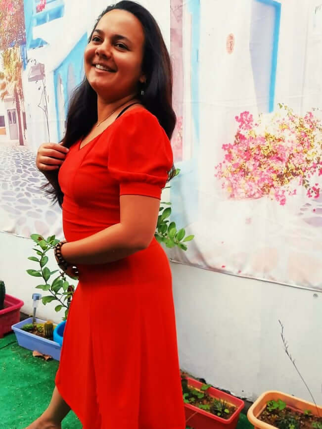 Femme souriante portant une robe rouge avec des manches ballons, devant un mur coloré et des plantes.
