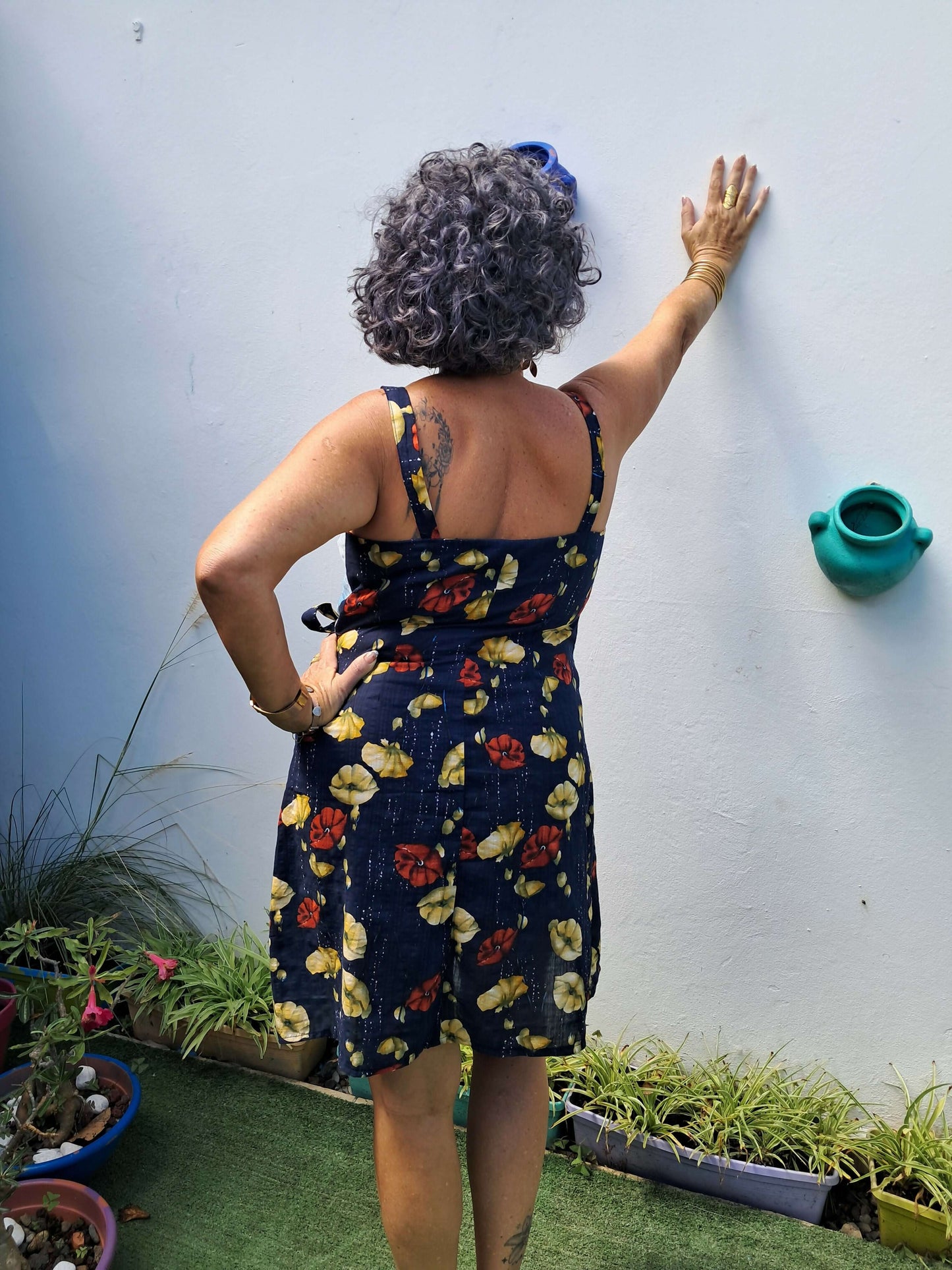 Femme portant la robe Ostara à bretelles, montrant un ourlet arrondi, dans un jardin avec pots de fleurs.