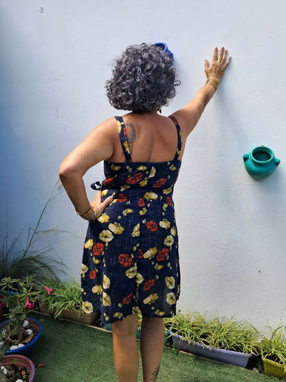 Femme portant la robe Ostara à bretelles, montrant un ourlet arrondi, dans un jardin avec pots de fleurs.