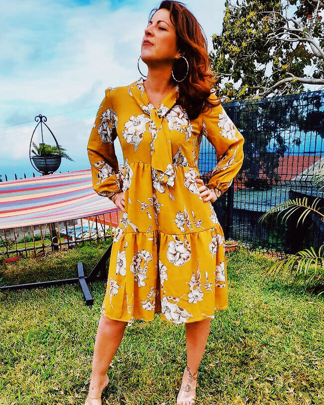 Femme portant une robe babydoll jaune à fleurs, style vintage avec un col Lavallière, dans un jardin.