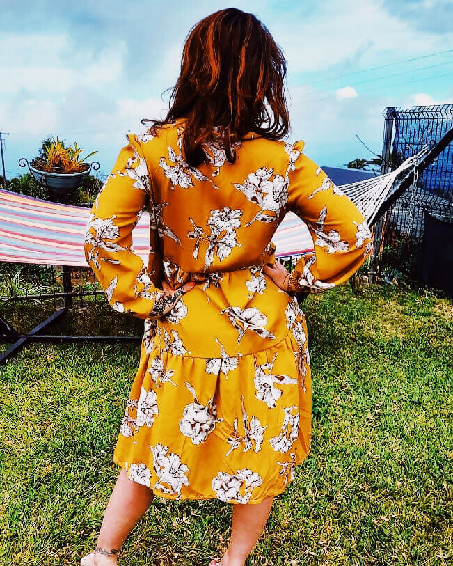 Femme portant une robe jaune à fleurs, vue de dos, dans un jardin avec un hamac en arrière-plan.