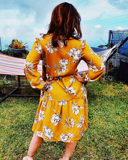 Femme portant une robe jaune à fleurs, vue de dos, dans un jardin avec un hamac en arrière-plan.