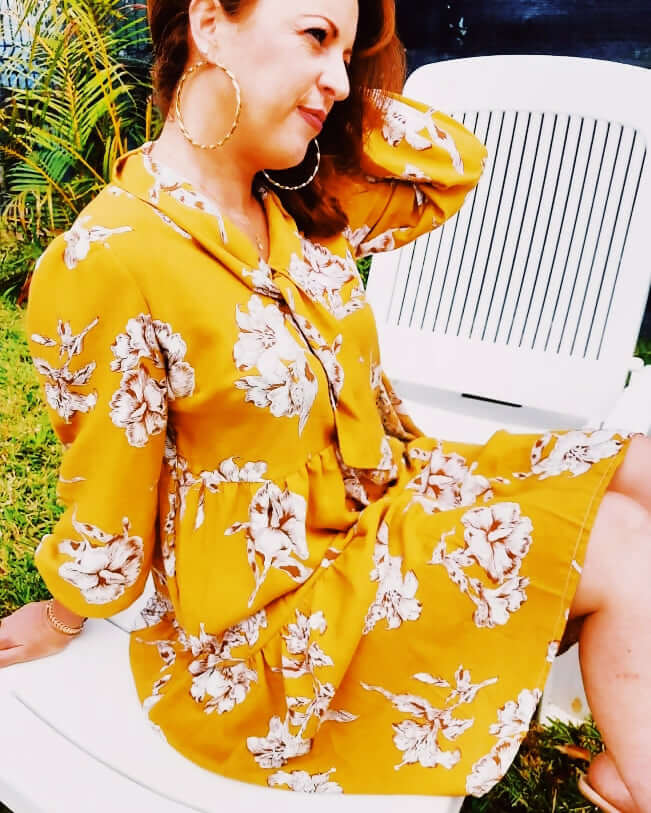 Femme souriante portant une robe babydoll jaune à fleurs, assise sur une chaise, ambiance décontractée en extérieur.