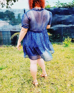 Femme portant une robe babydoll en dentelle bleu, vue de dos, dans un jardin verdoyant.