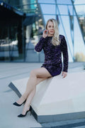 Femme en robe pailletée violette et chaussures noires, assise sur un banc moderne.
