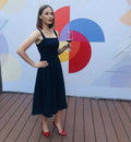 Femme portant une robe élégante, debout sur une terrasse en bois, tenant un verre de vin rouge, mur coloré en arrière-plan.