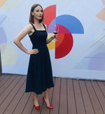 Femme portant une robe élégante, debout sur une terrasse en bois, tenant un verre de vin rouge, mur coloré en arrière-plan.