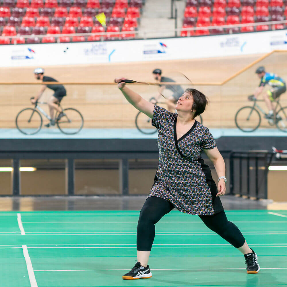 Femme en mouvement portant la robe tunique sportwear Cranberry, jouant au badminton, sur fond de cyclistes en indoor.