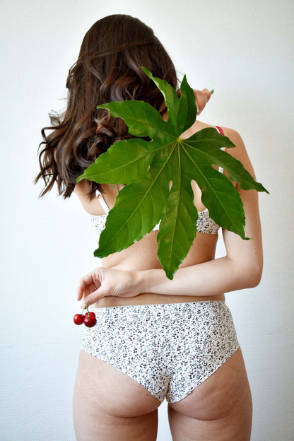 Femme de dos avec un maillot de bain floral tenant des cerises, cachée par une grande feuille.