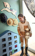 Enfant jouant avec un cheval en bois, devant une maison de poupée, portant un costume de cavalier beige et blanc.