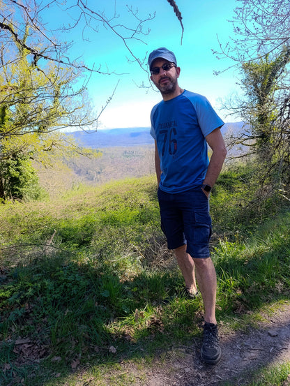 Homme en t-shirt bleu et shorts, posant dans un paysage verdoyant sous un ciel clair.