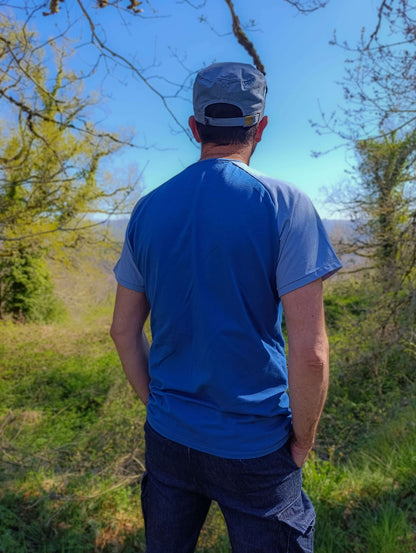 Homme de dos portant un t-shirt bleu en plein air, dans un environnement naturel verdoyant.