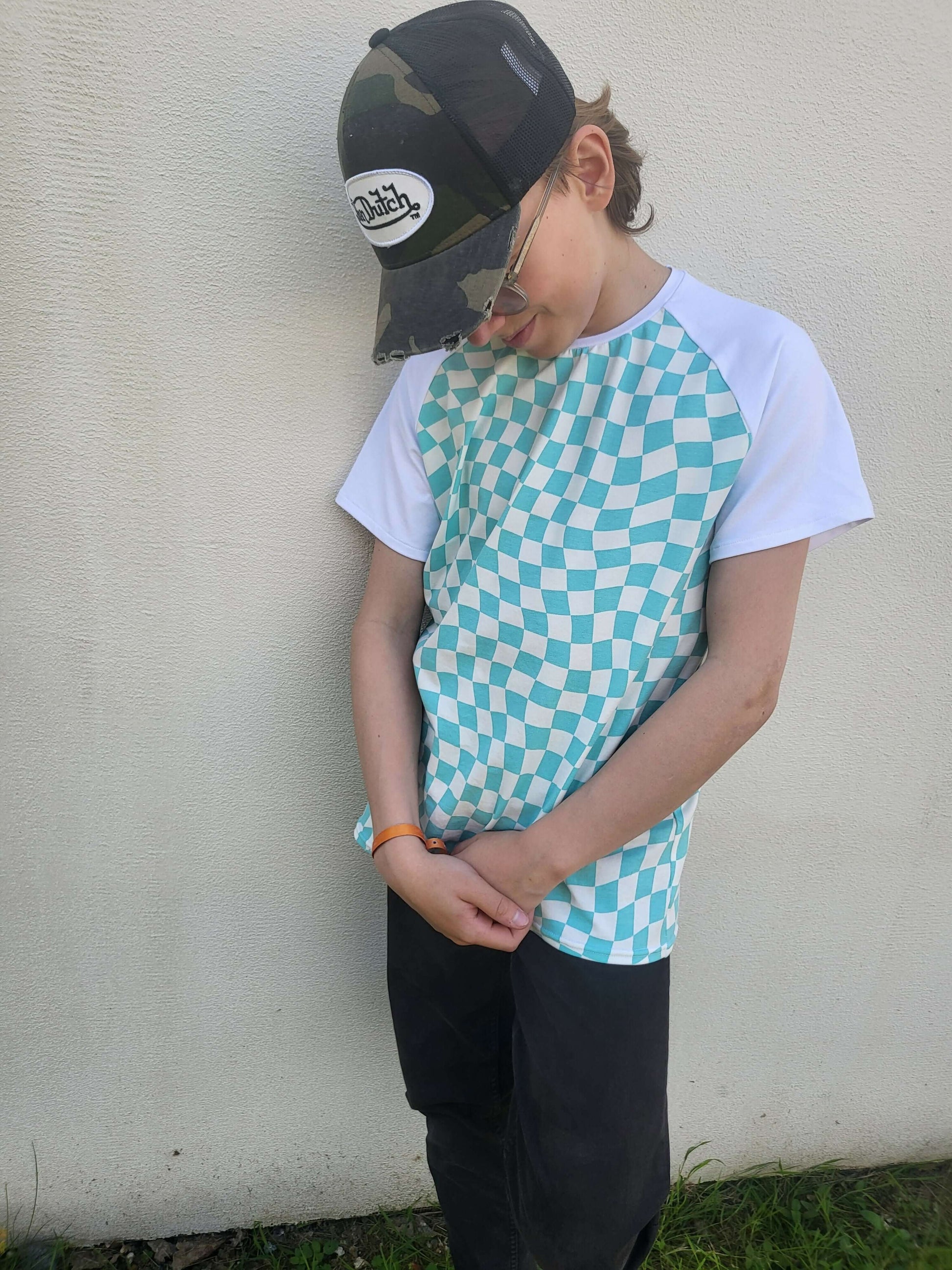 Enfant portant un t-shirt à motif en damier turquoise et blanc, accompagné d'une casquette camouflée.