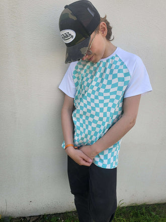 Enfant portant un t-shirt à motif en damier turquoise et blanc, accompagné d'une casquette camouflée.