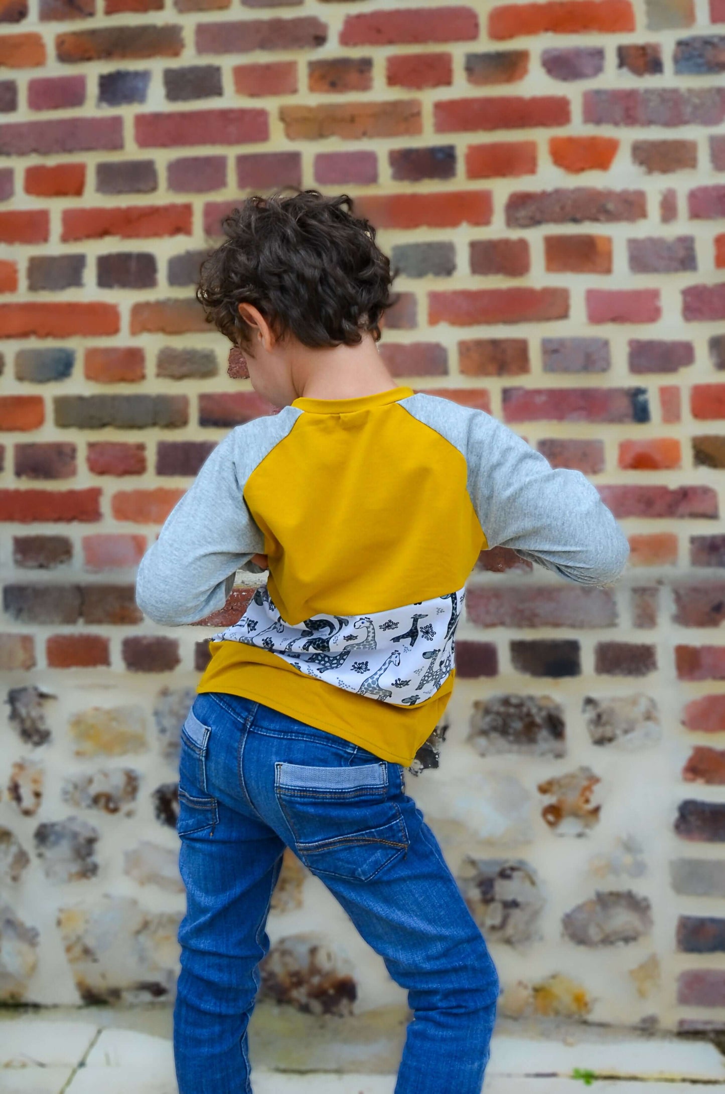 Enfant portant un t-shirt Loulou avec manches raglan, affichant un style unique et tendance devant un mur en brique.
