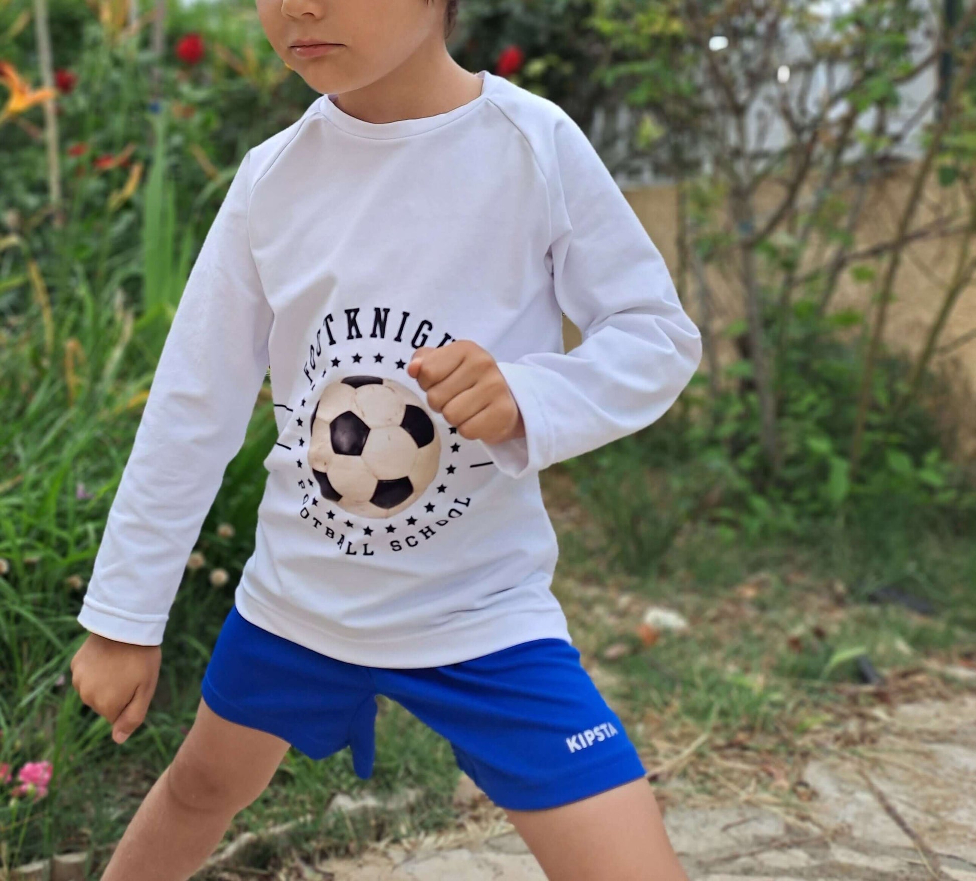 Enfant en t-shirt blanc avec motif football, jouant dehors sur pelouse, et portant un short bleu. 