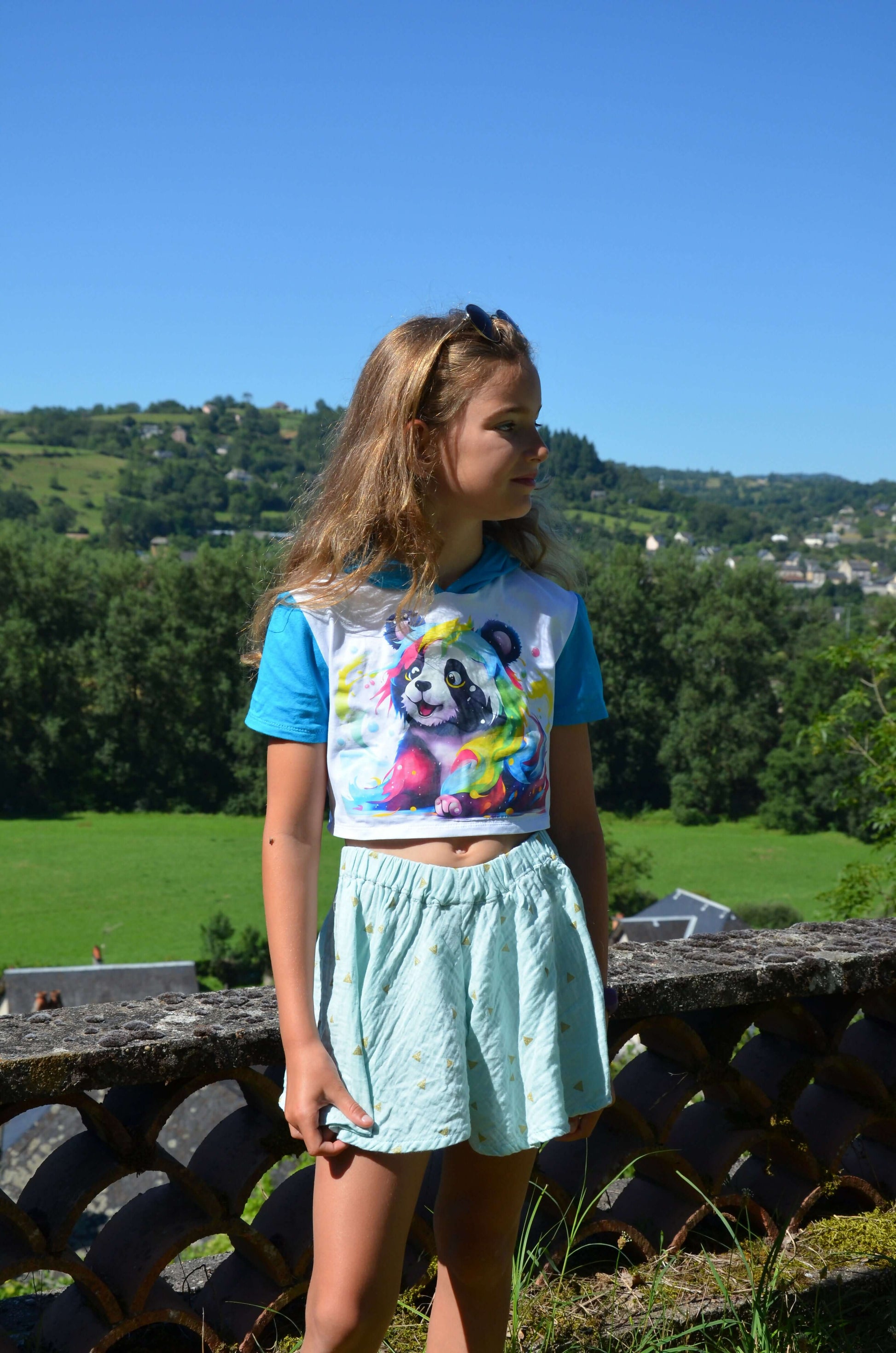 Fille portant un t-shirt bleu avec un panda coloré et une jupe courte, devant un paysage verdoyant.