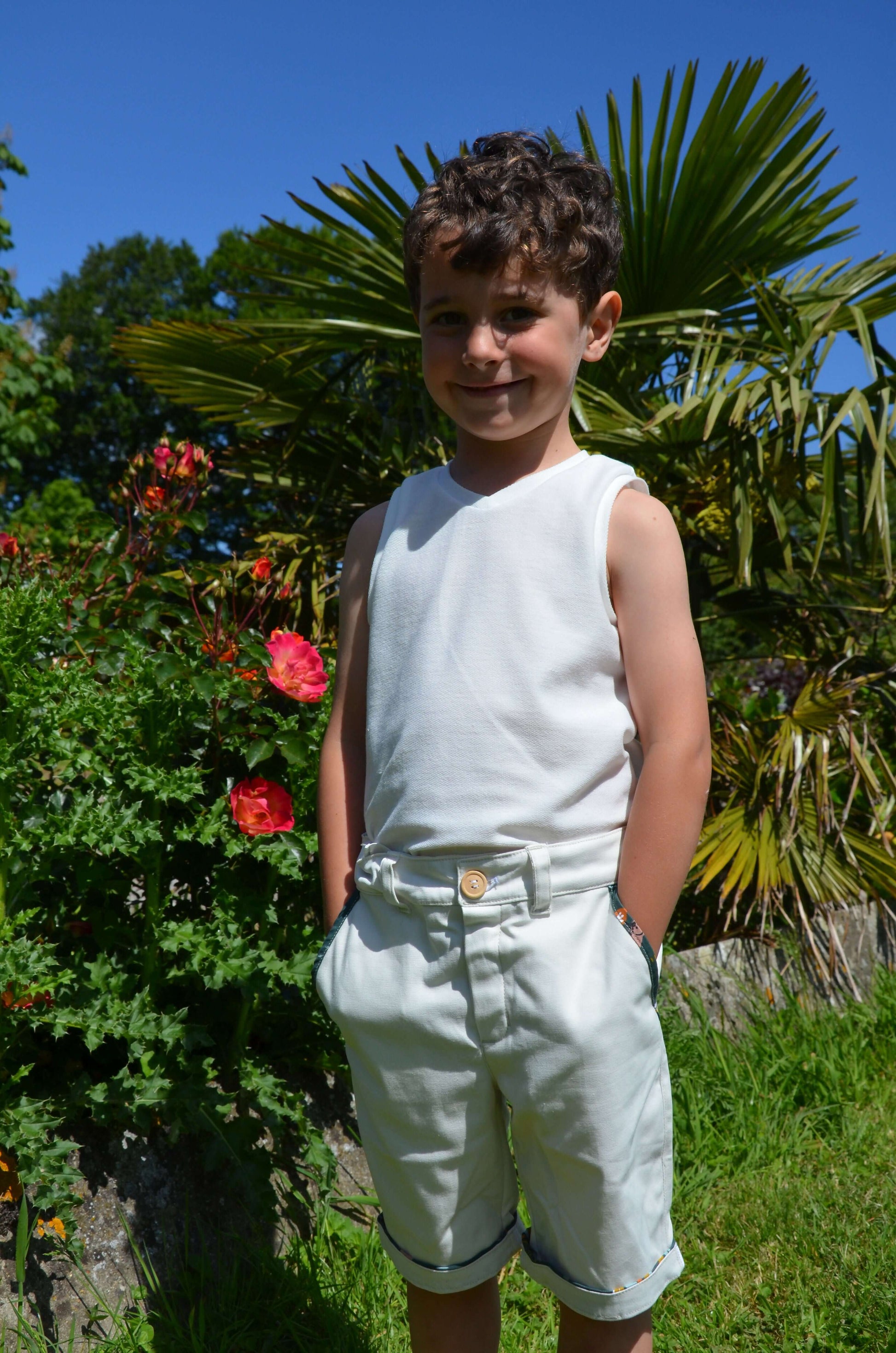 Un enfant souriant en débardeur blanc et short beige, en pleine nature, avec des fleurs en arrière-plan.