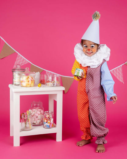 Déguisement Analya le Clown : enfant en costume coloré avec chapeau, sourire, devant une table de bonbons.