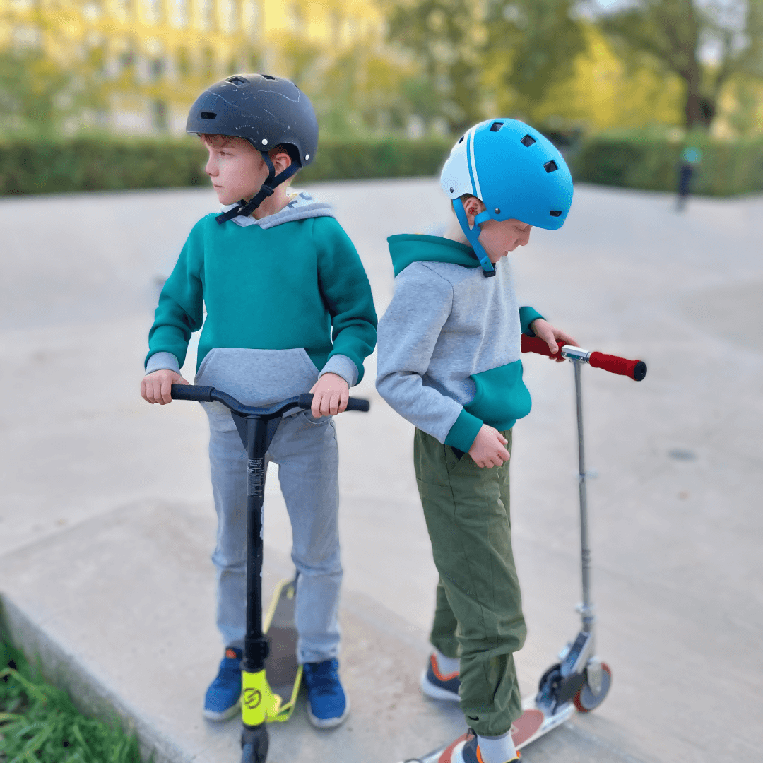 Deux garçons en sweats Calao et casques, jouant avec des trottinettes au skatepark.