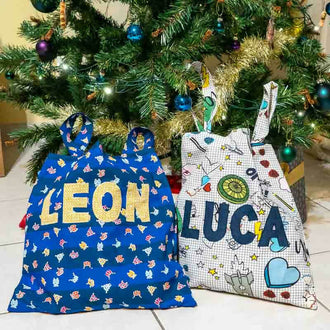 Sacs personnalisés avec lettres appliquées, décorés pour Noël, posés sous un arbre de Noël.