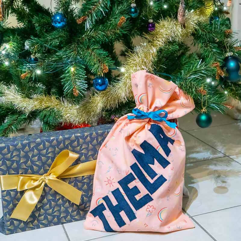 Sac décoré du prénom THELMA avec appliqués de lettres, sous un sapin de Noël.