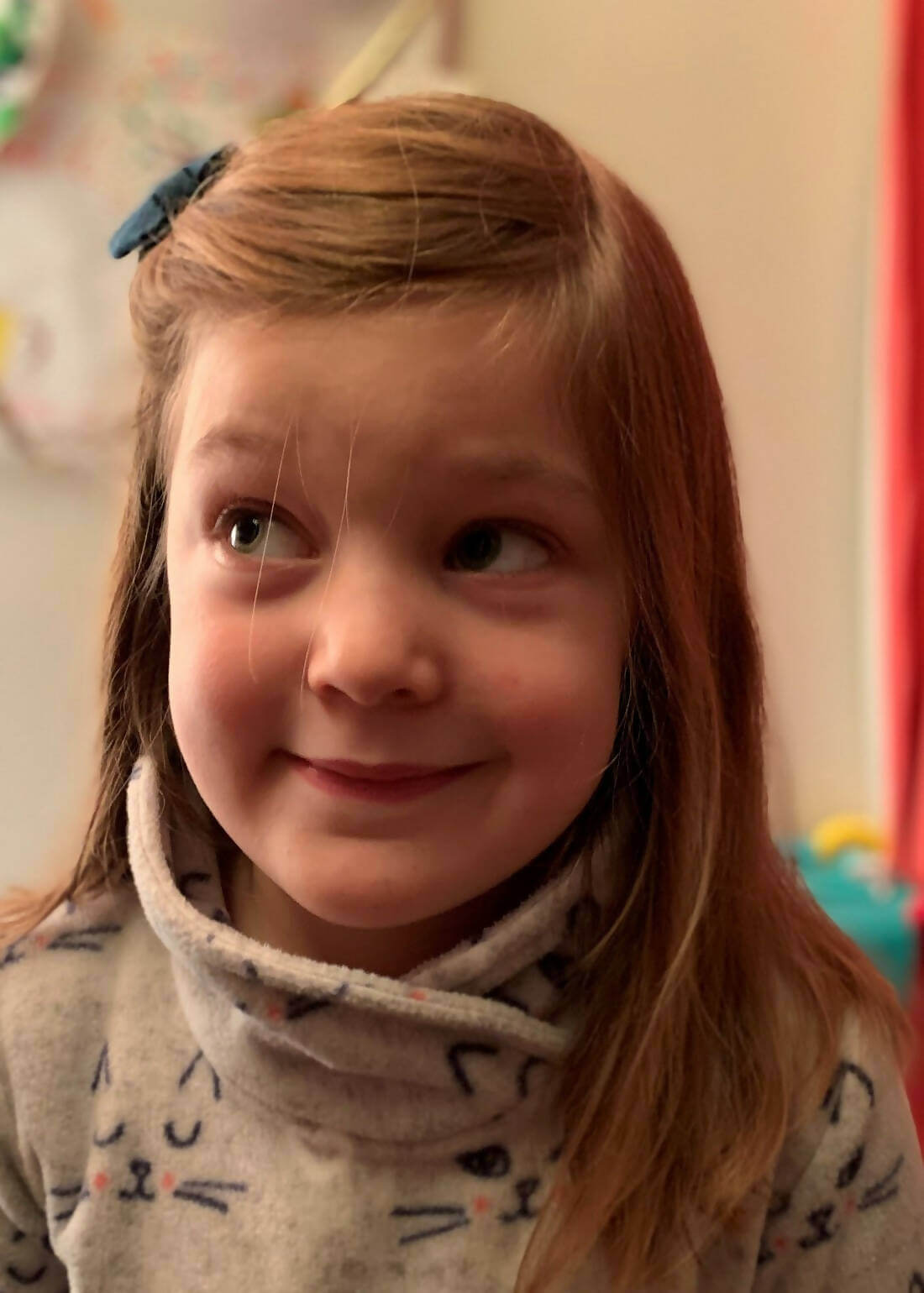 Enfant portant une robe-sweat avec motif de chat et col roulé, sourire espiègle et barrette bleue dans les cheveux, dans une pièce colorée.