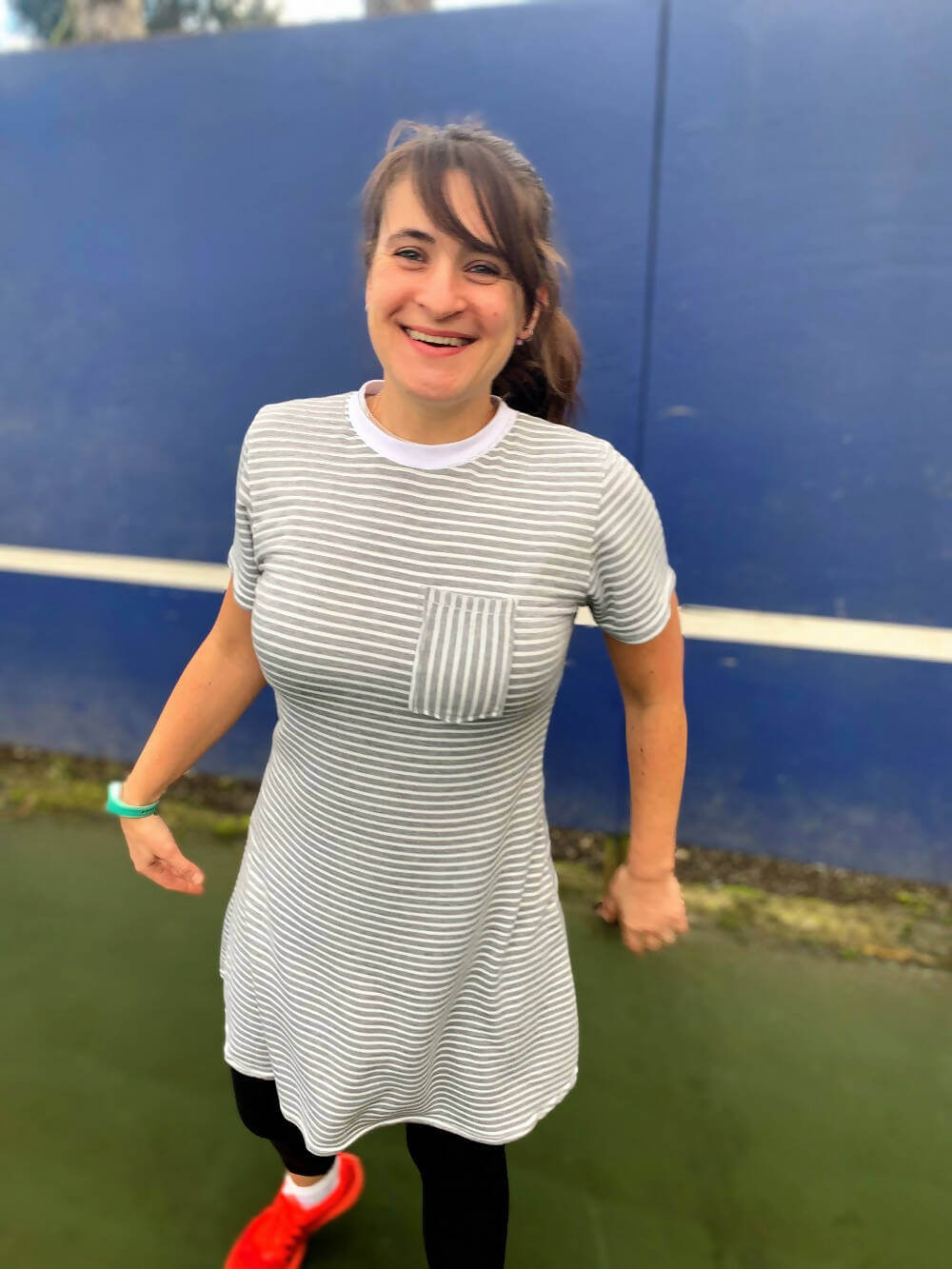 Femme souriante portant une robe rayée grise et blanche avec poche avant, sur un terrain de sport.