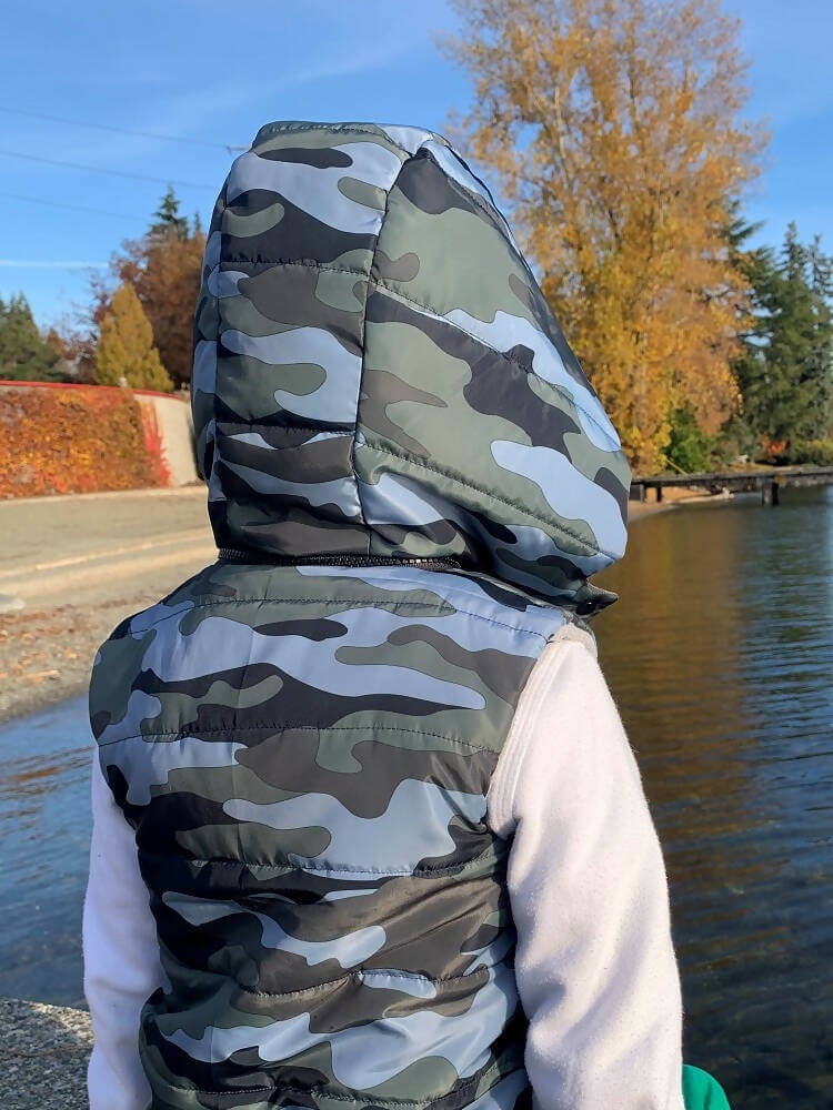 Enfant portant une Mini-KEZIA - Doudoune sans manches mixte pour enfants du 2 au 16 Ans, motif camouflage et capuche amovible.