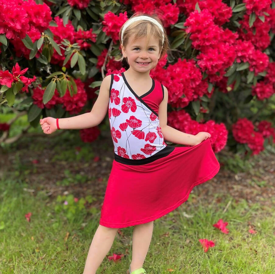 Fillette portant la Mini-SANDY - Robe cache-coeur pour filles du 2 au 16 Ans, devant un buisson fleuri. Patron de couture pdf à télécharger disponible.