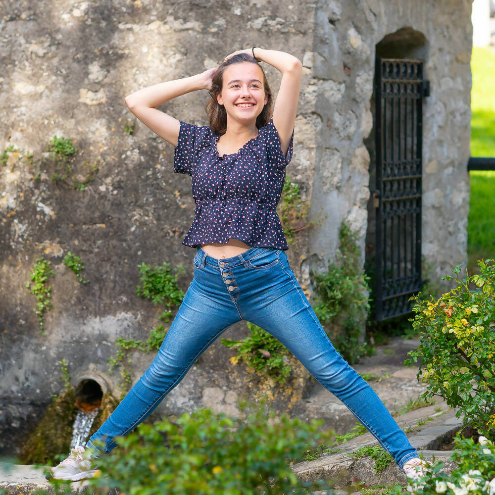 Jeune femme souriante portant un crop top fleuri et un jean, devant un mur de pierre avec végétation.