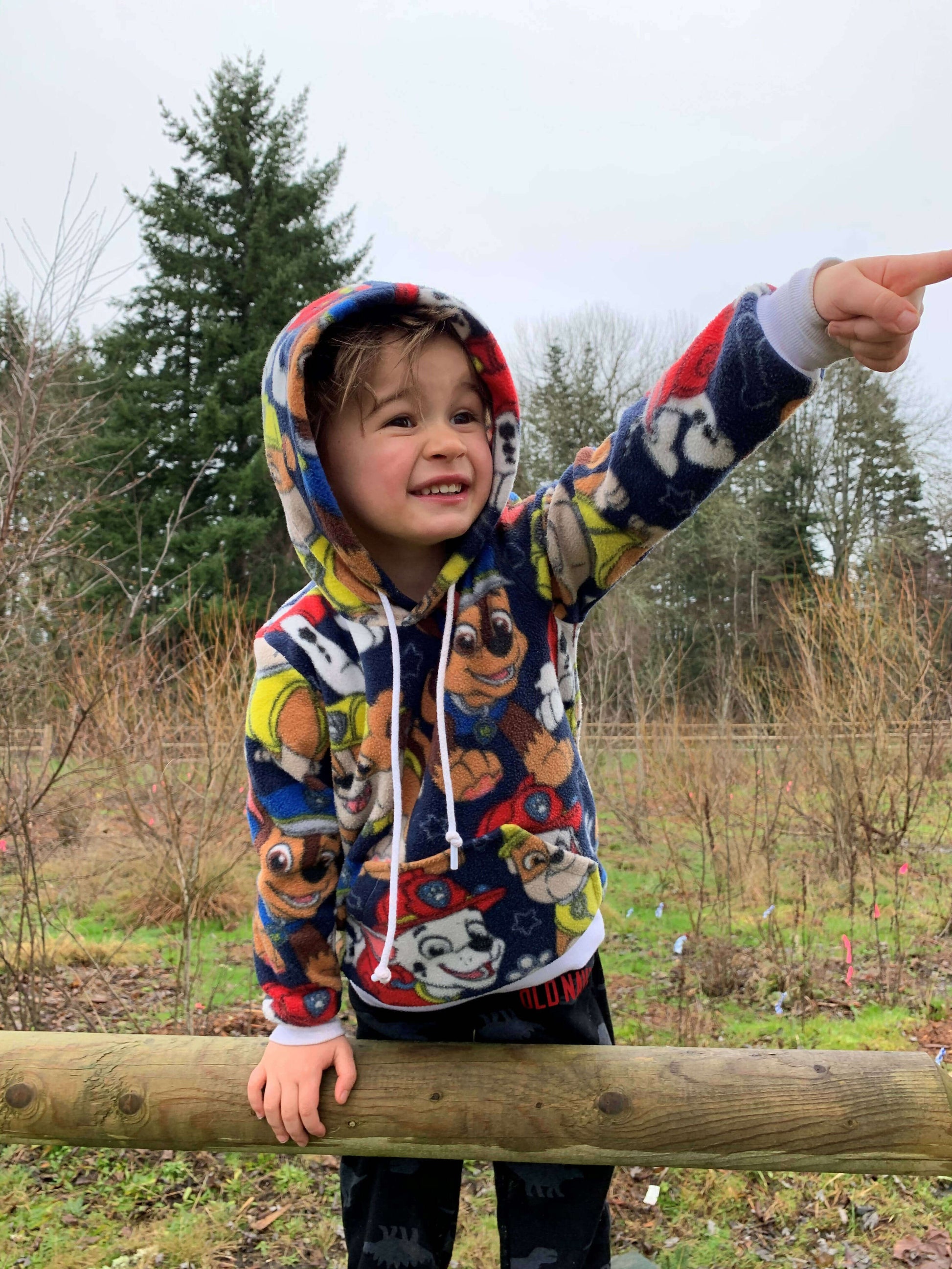 Enfant souriant portant un sweat à capuche coloré, pointant vers l'horizon lors d'une promenade en plein air.
