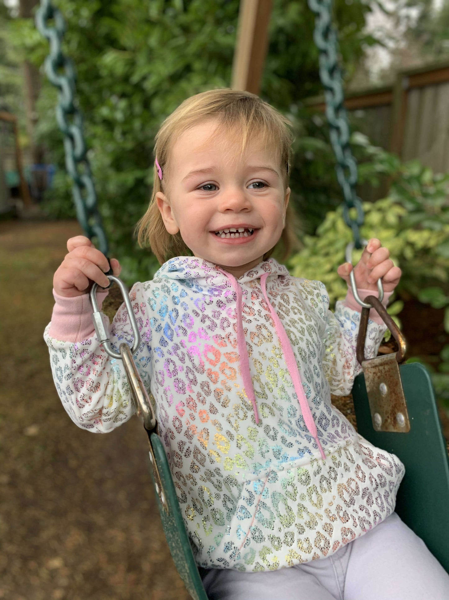 Enfant souriant portant un sweat à imprimé léopard, assise sur une balançoire dans un jardin verdoyant.